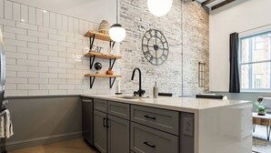 A modern kitchen with white subway tiles, a brick accent wall, a large clock, and a minimalist design. - Cobblestone Chic - WiFi/Downtown/Walk Score 91 (Birmingham)
