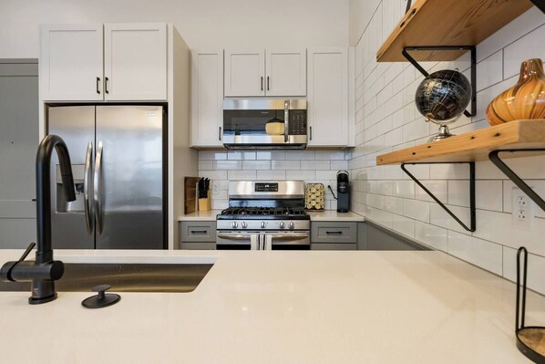 Una cocina moderna con armarios blancos, electrodomésticos de acero inoxidable y un estante de madera con objetos decorativos.