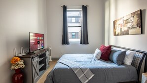 A bedroom with a bed, a TV, a desk, a vase with flowers, and a window with curtains. - Cobblestone Chic - WiFi/Downtown/Walk Score 91 (Birmingham)