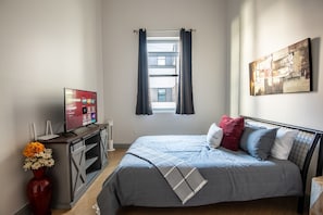 A bedroom with a bed, a TV, a desk, a vase with flowers, and a window with curtains.