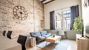 A modern living room with a brick wall, a large round clock, a grey sofa, a wooden coffee table, and a TV on a wooden stand. - Cobblestone Chic - WiFi/Downtown/Walk Score 91 (Birmingham)