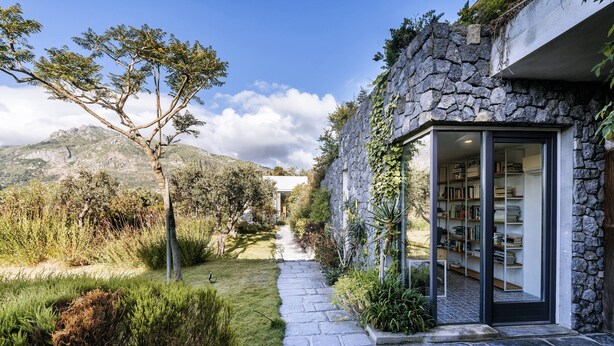 A stone house with a stone exterior, a glass door leading to a library, and a pathway leading to a mountain view.