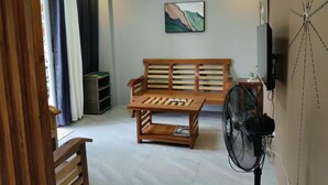 Una sala de estar con un sofá de madera, una mesa de centro, un televisor montado en la pared y un ventilador de pie.