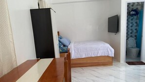 Ein Zimmer mit einem hölzernen Bett, einem an der Wand befestigten Fernseher, einer weißen Tür und einem durch eine geöffnete Tür einsehbaren Badezimmer.