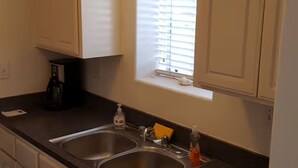 A kitchen with white cabinets, a double sink, a window with blinds, and a ceiling light fixture. - SC Cozy Beach Apartment (San Clemente)