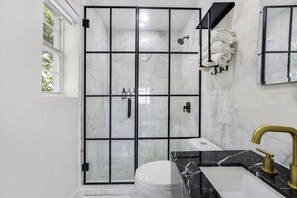 A modern bathroom with a glass shower enclosure, a white toilet, a black marble sink, and a gold faucet.