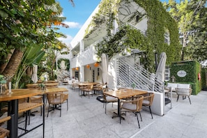 An outdoor café with wooden tables and wicker chairs, surrounded by greenery and a white building with a staircase. - Lemon Branch (Miami Beach)
