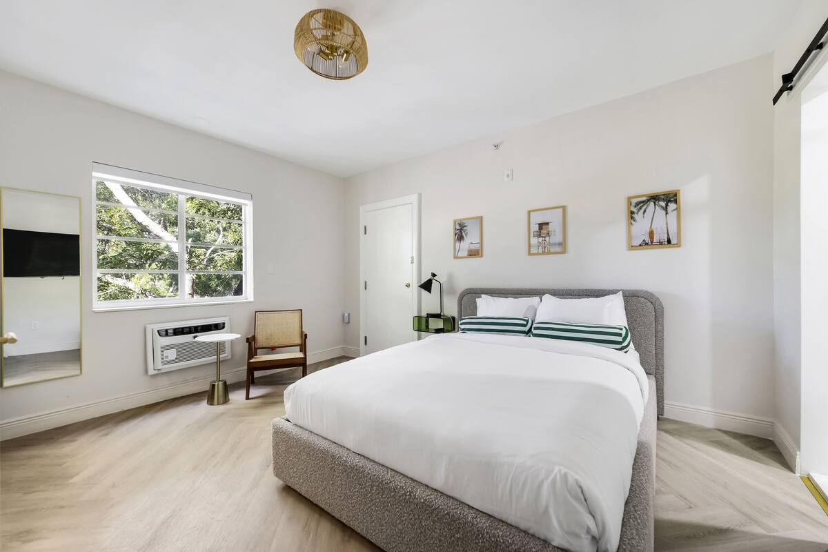 A bedroom with a large bed, a window, a chair, a bedside table, and a wall-mounted TV.