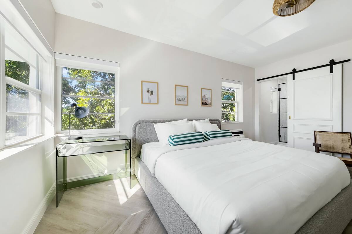 A bedroom with a bed, a glass desk, and a window with a view of trees.