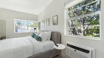 A bedroom with a bed, bedside table, air conditioning unit, and large windows with a view of trees.
