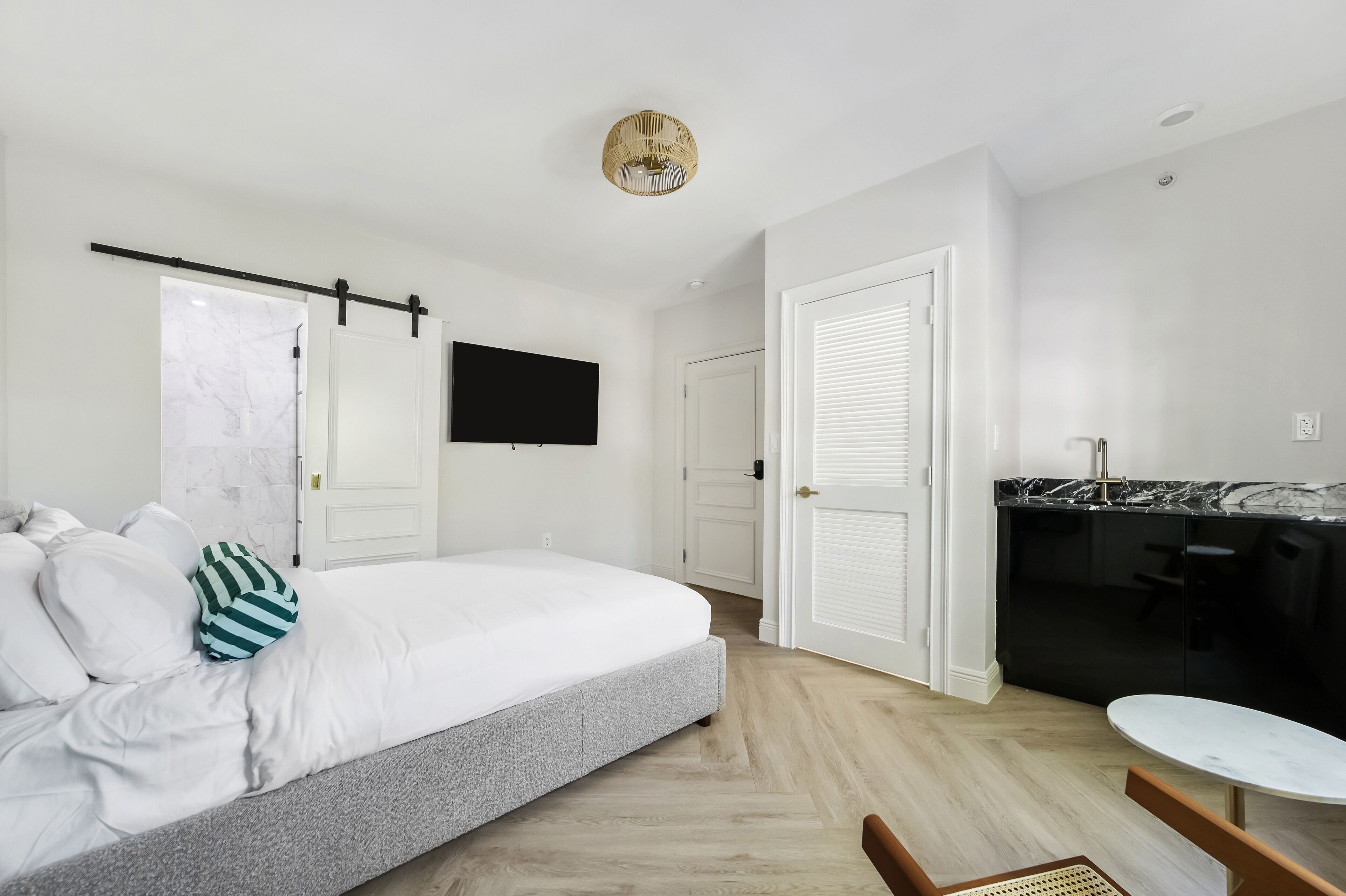 Quarto com cama, televisão, porta e área de cozinha com bancada e cadeiras.
