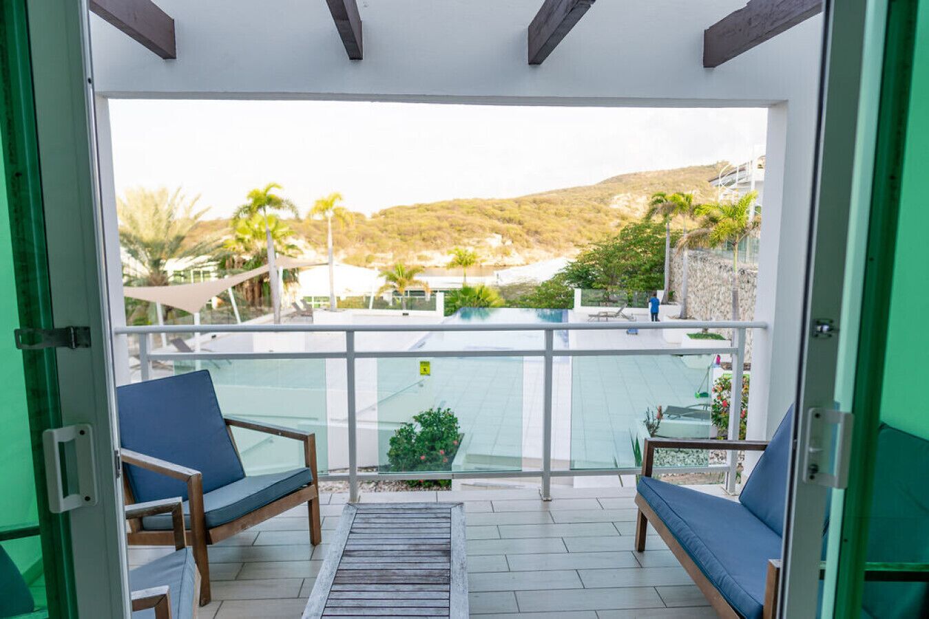 A balcony with two blue cushioned chairs, a small table, and a view of a pool and surrounding landscape.