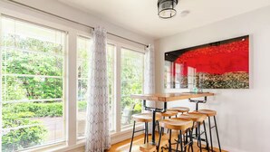 A modern kitchen with a wooden bar and stools, large windows with blinds, and a red abstract painting on the wall.