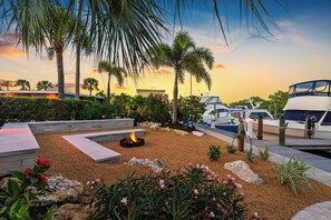 A landscaped outdoor area with a fire pit, palm trees, and boats docked at a marina.