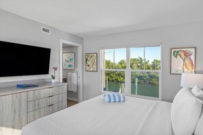 A bedroom with a large bed, a flat-screen TV, a window with a view of a body of water, and artwork on the walls.