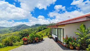 Un edificio moderno con techo rojo y ventanas amplias, rodeado de abundante vegetación y un paisaje montañoso.