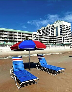 Une plage avec deux chaises longues et un parasol rouge et bleu. En arrière-plan, on aperçoit un grand bâtiment d’hôtel avec de nombreux étages et des balcons.