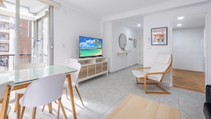 Un moderno salón con una mesa de vidrio, sillas blancas, un televisor sobre un mueble de madera, un sofá blanco y suelo de madera.