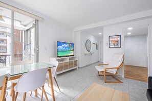 A modern living room with a glass dining table, white chairs, a TV on a wooden stand, a white sofa, and a wooden floor. - Casablanca 2-D Apartment Levante Beach (Benidorm)