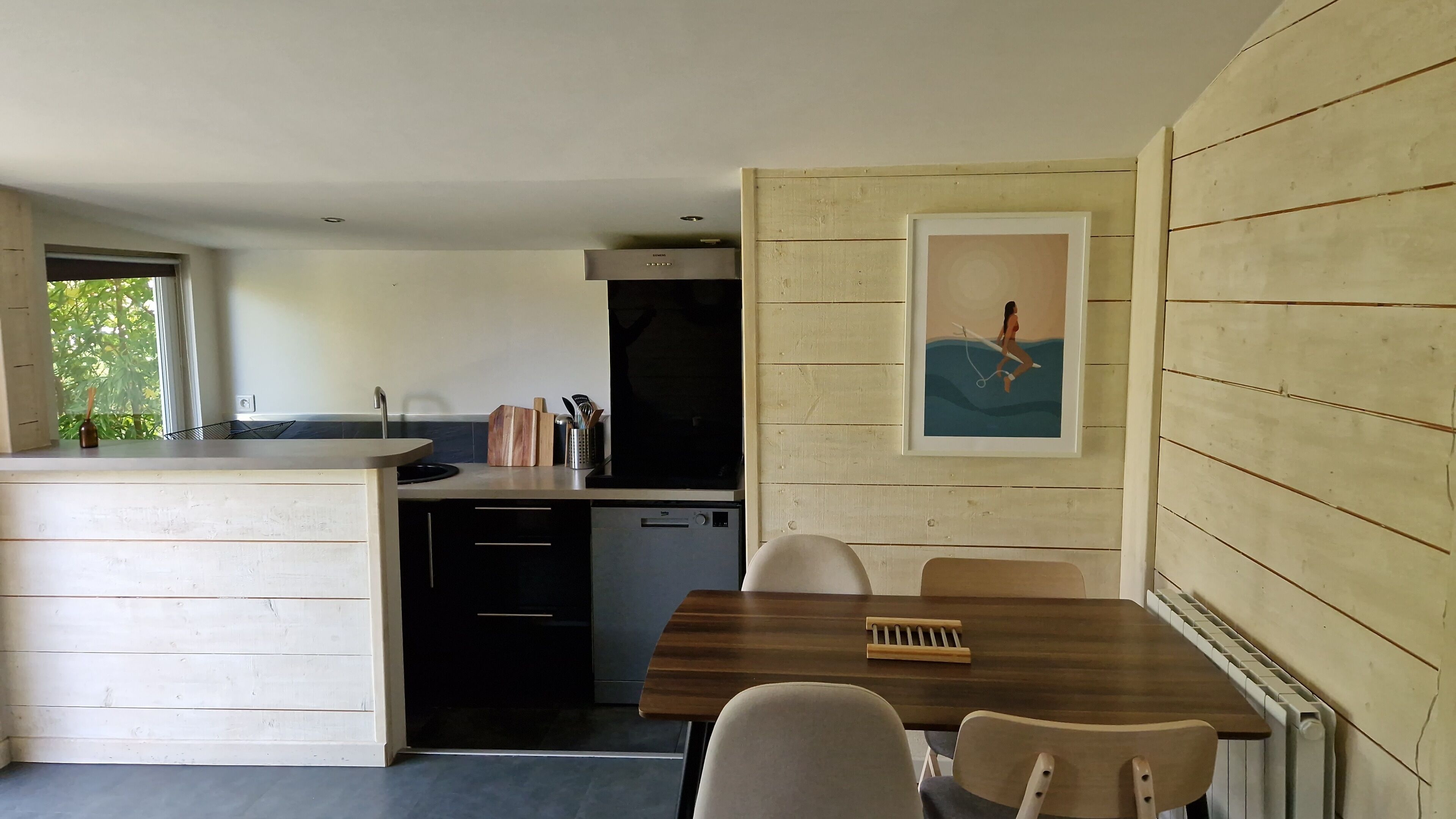 A compact kitchen with a built-in dishwasher, a wooden dining table with chairs, and a framed picture on the wall.