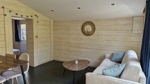 Une pièce chaleureuse avec un mur en bois, une table ronde éclairée par une bougie, un canapé blanc agrémenté de coussins à motifs et une table à manger en bois avec des chaises.