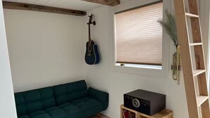 Une pièce chaleureuse avec un canapé vert, une guitare, une radio et une fenêtre équipée de stores.