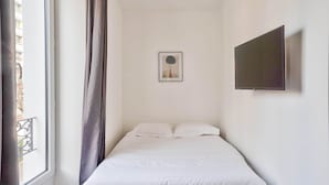 Een slaapkamer met een bed, een raam met gordijnen, een lijst met een afbeelding aan de muur, en een flatscreen televisie die aan de muur is bevestigd.