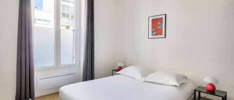 A bedroom with a bed, a nightstand, a red lamp, a white pillow, a framed picture on the wall, and a window with curtains.