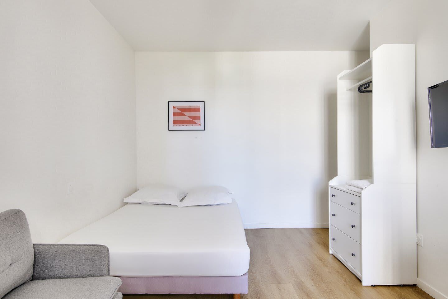 Une chambre à coucher au style minimaliste, avec un lit, un fauteuil gris, une commode et un tableau encadré au mur.