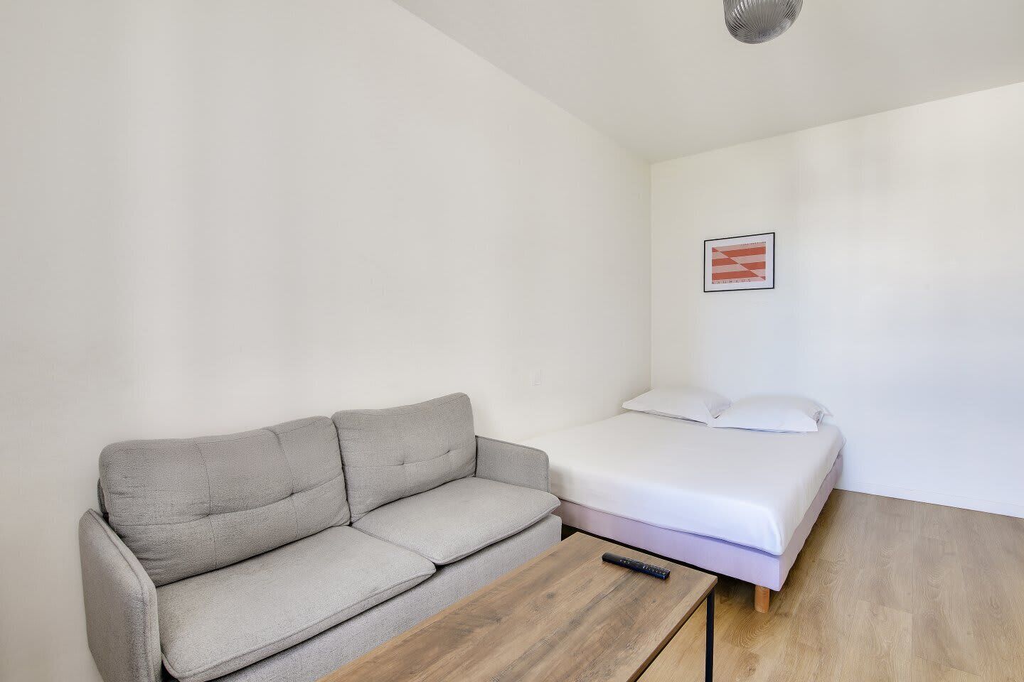 A small, minimalist room with a grey sofa, a single bed, and a wooden table.