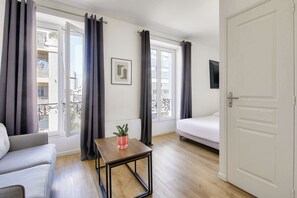 A modern room with a grey sofa, a small wooden table with a plant, a white bed, and a white door. - Beautifully furnished cosy studio with a street view (Ivry-sur-Seine)