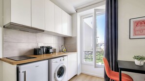 A compact kitchen with a washing machine, a wooden countertop, and a dining area with a chair and table. - Beautifully furnished cosy studio with a street view (Ivry-sur-Seine)