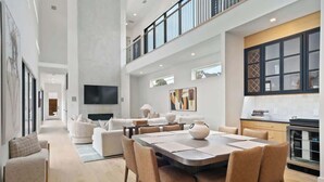 A spacious living area with a high ceiling, a dining table, sofas, and a kitchen with a large window. - Lux 4100sqft Estate•Rooftop•Pool/Spa (Dallas)