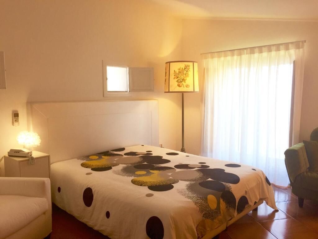 Hotellihuone, jossa on sänky, yöpöytä, lamppu, tuoli ja ikkuna, jossa on verhot.
