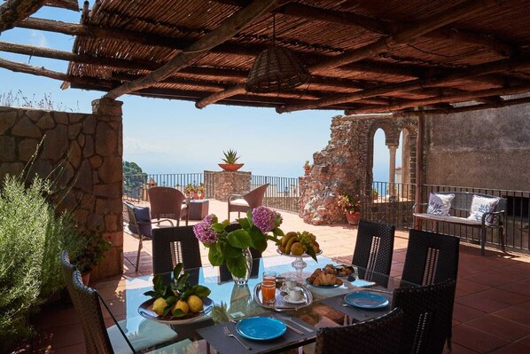 An outdoor dining area with stone walls, wicker furniture, and a view of the sea.