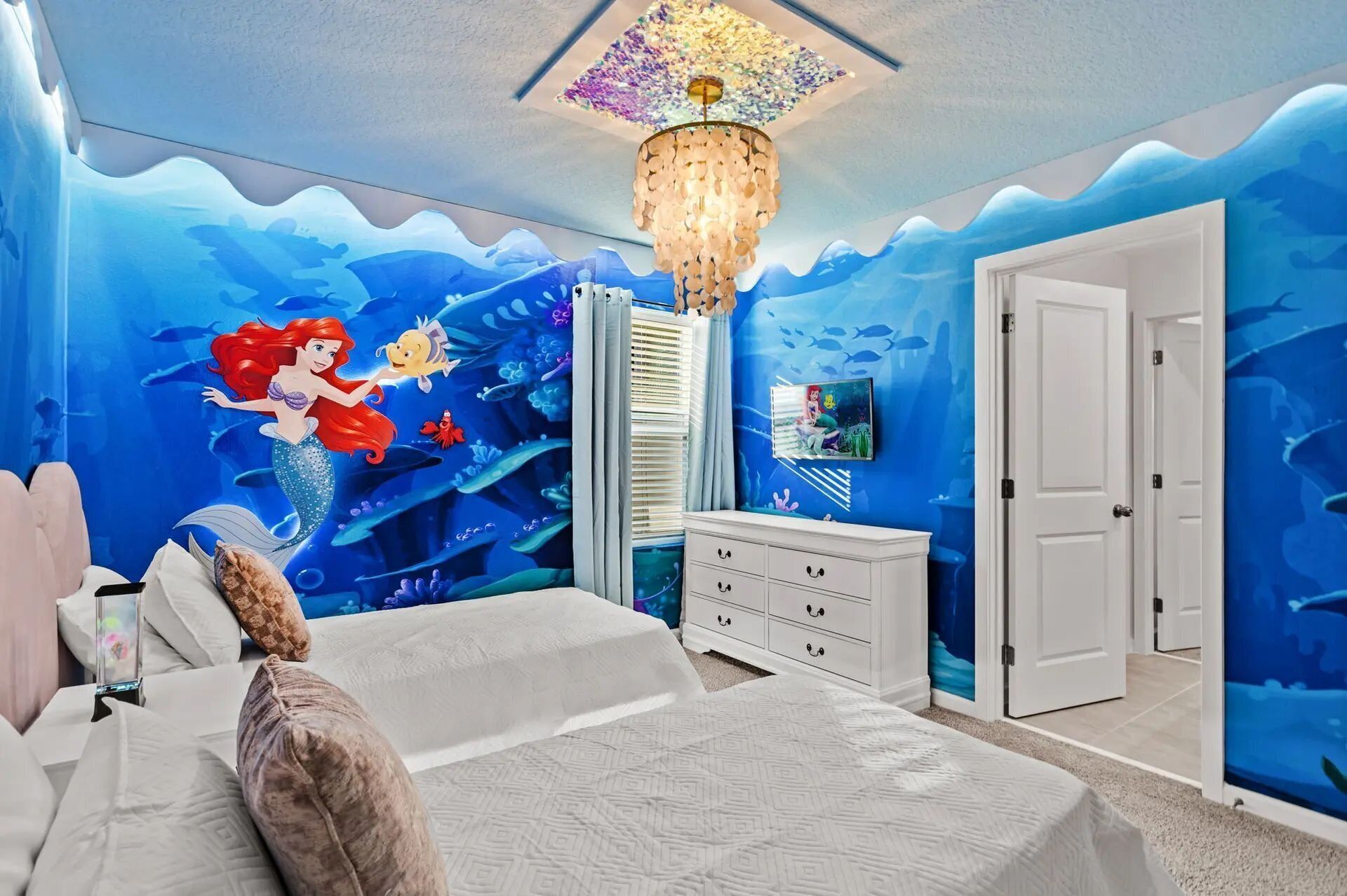 Une chambre à coucher avec une fresque représentant une sirène, un lustre, un lit, une commode et une porte.