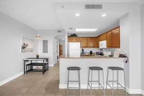 A modern kitchen with wooden cabinets, a central island with bar stools, a dining area with a table and chairs, and a pendant light. - Renovated & Exclusive Lake Condo w/Disney view! (Orlando)