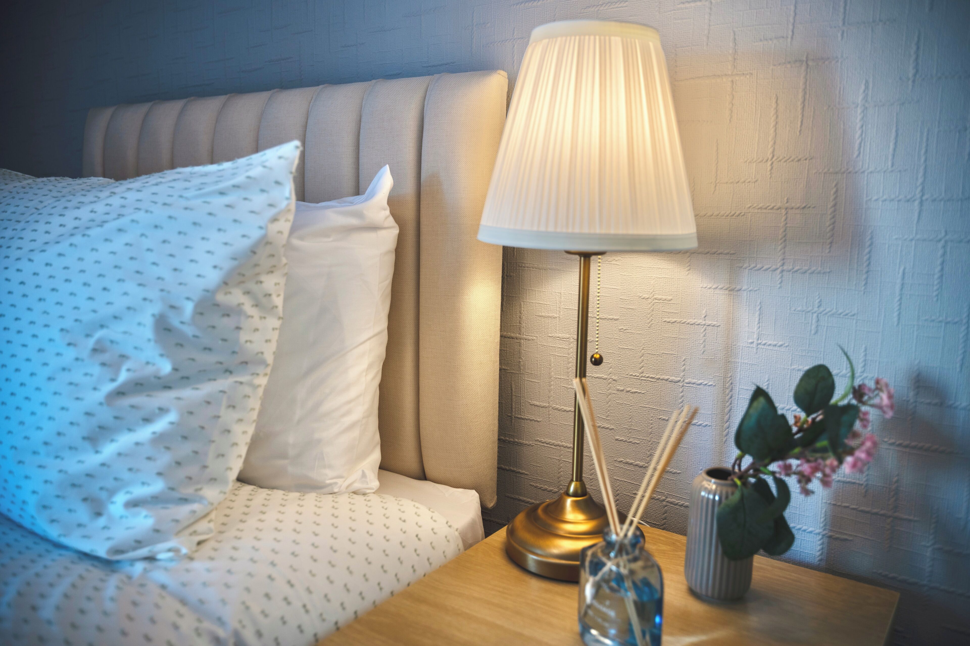 A neatly made bed with a patterned bedspread and pillows, a bedside lamp, a small vase with flowers, and a textured wall behind.