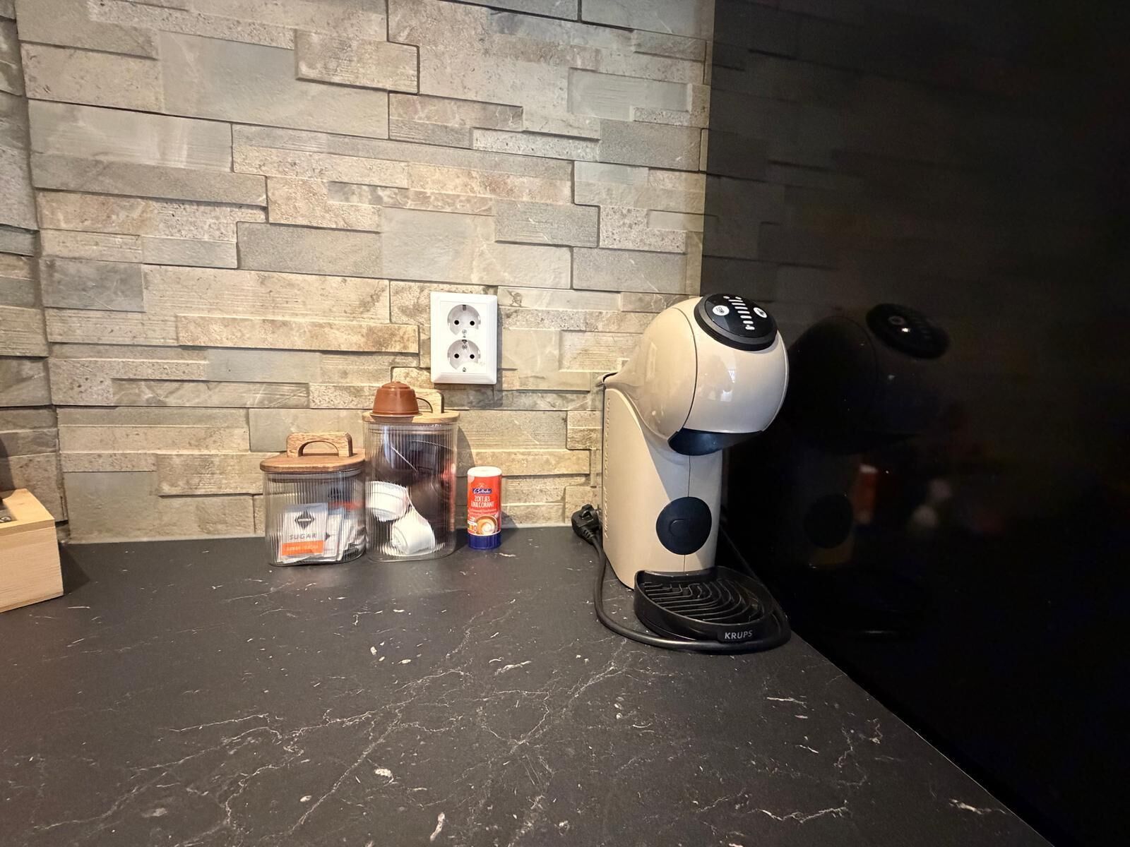Una cocina con pared de piedra, una cafetera, un tarro de azúcar, un envase de crema para café y una pequeña caja de madera.