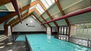 An indoor swimming pool with a high ceiling, wooden beams, and a logo on the wall. - VELUWE WELLNESS CHALET - Private Hot Tub and Sauna on Park with Swimming Pool (Voorthuizen)