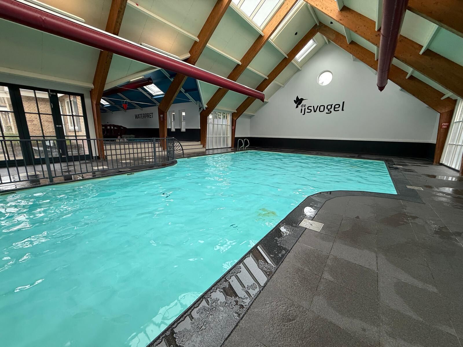 An indoor swimming pool with a high ceiling and wooden beams, a 'WATERPARK' sign, and a logo for 'de ijsvogel'.