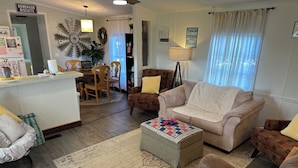 Ein gemütliches Wohnzimmer mit einem beigen Sofa, einem gemusterten Ottomanen und einem Couchtisch. Im Hintergrund ist ein Essbereich mit einer Kristallleuchter und einem Schild mit der Aufschrift "Welcome" zu sehen.