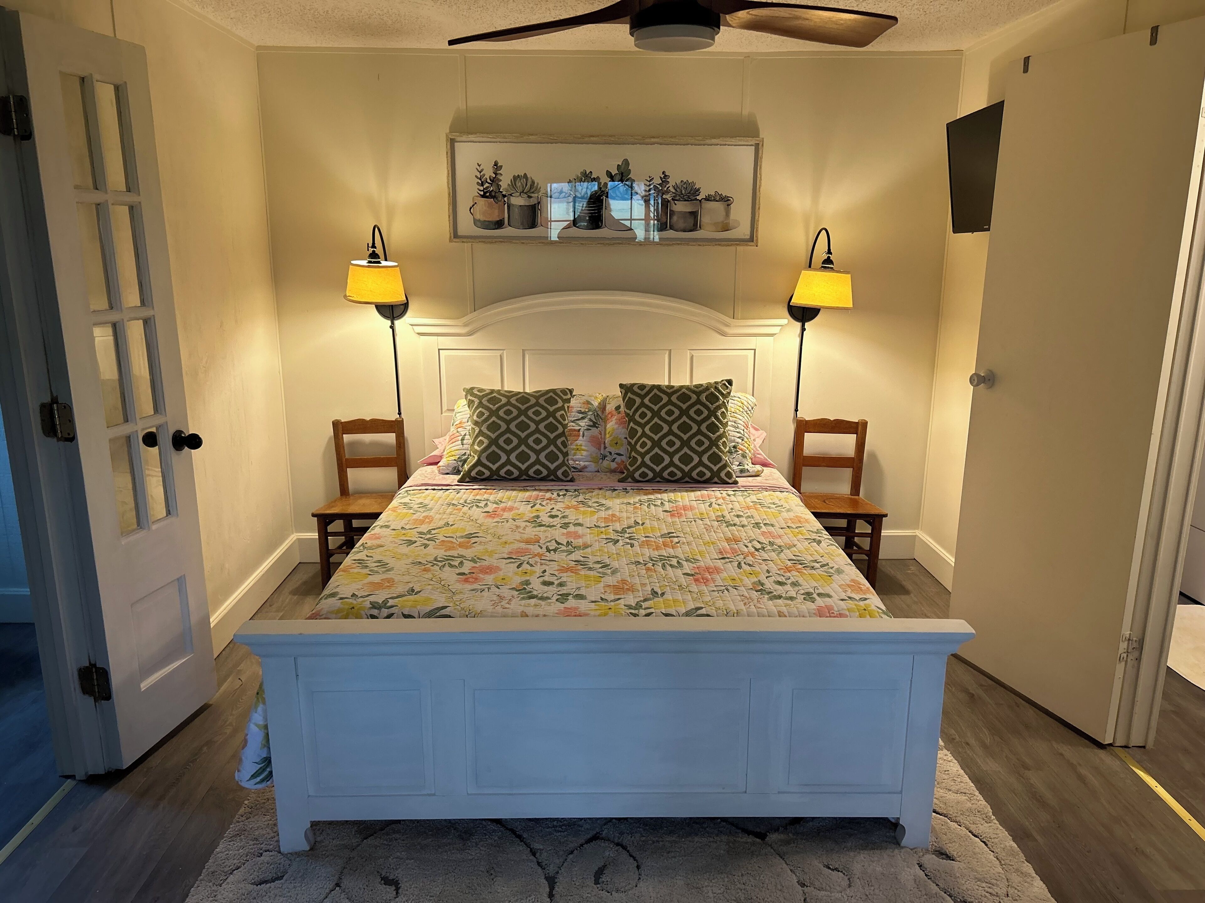 A bedroom with a bed, two chairs, a nightstand, a lamp, a ceiling fan, and a wall-mounted shelf with potted plants.