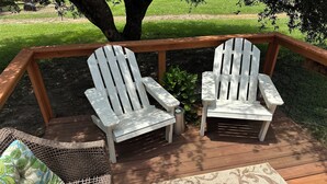 Eine Terrasse mit zwei Adirondack-Stühlen, einem Korbstuhl und einem gemusterten Teppich.