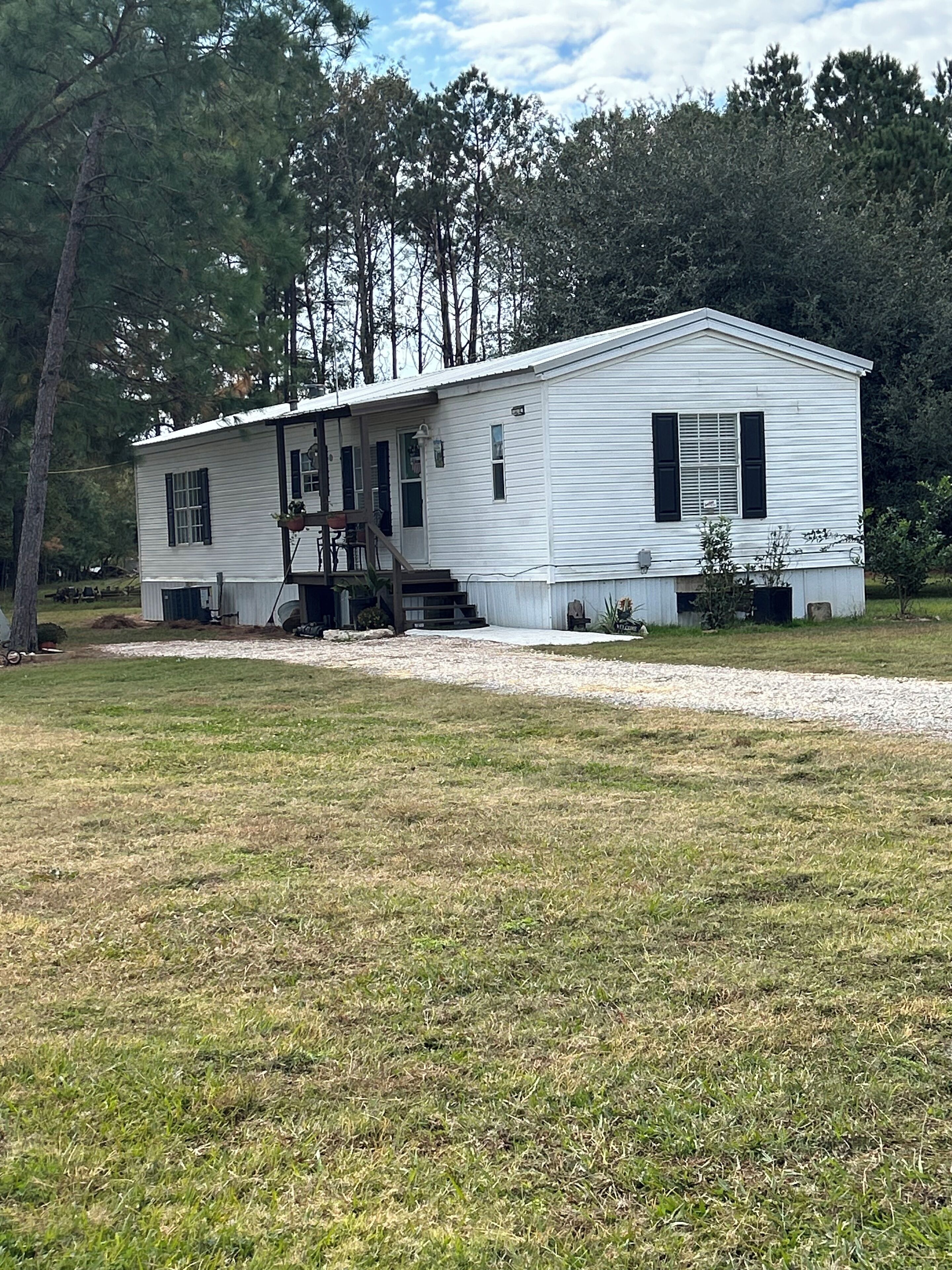A single-story mobile home with a front porch and a small deck, surrounded by a grassy yard and trees.
