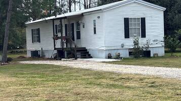 A single-story mobile home with a front porch and a small deck, surrounded by a grassy yard and trees.
