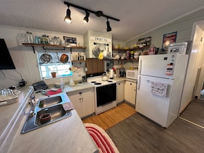 Cocina compacta con lavabo, estufa, microondas y refrigerador. Hay repisas sobre la encimera con frascos y utensilios de cocina.