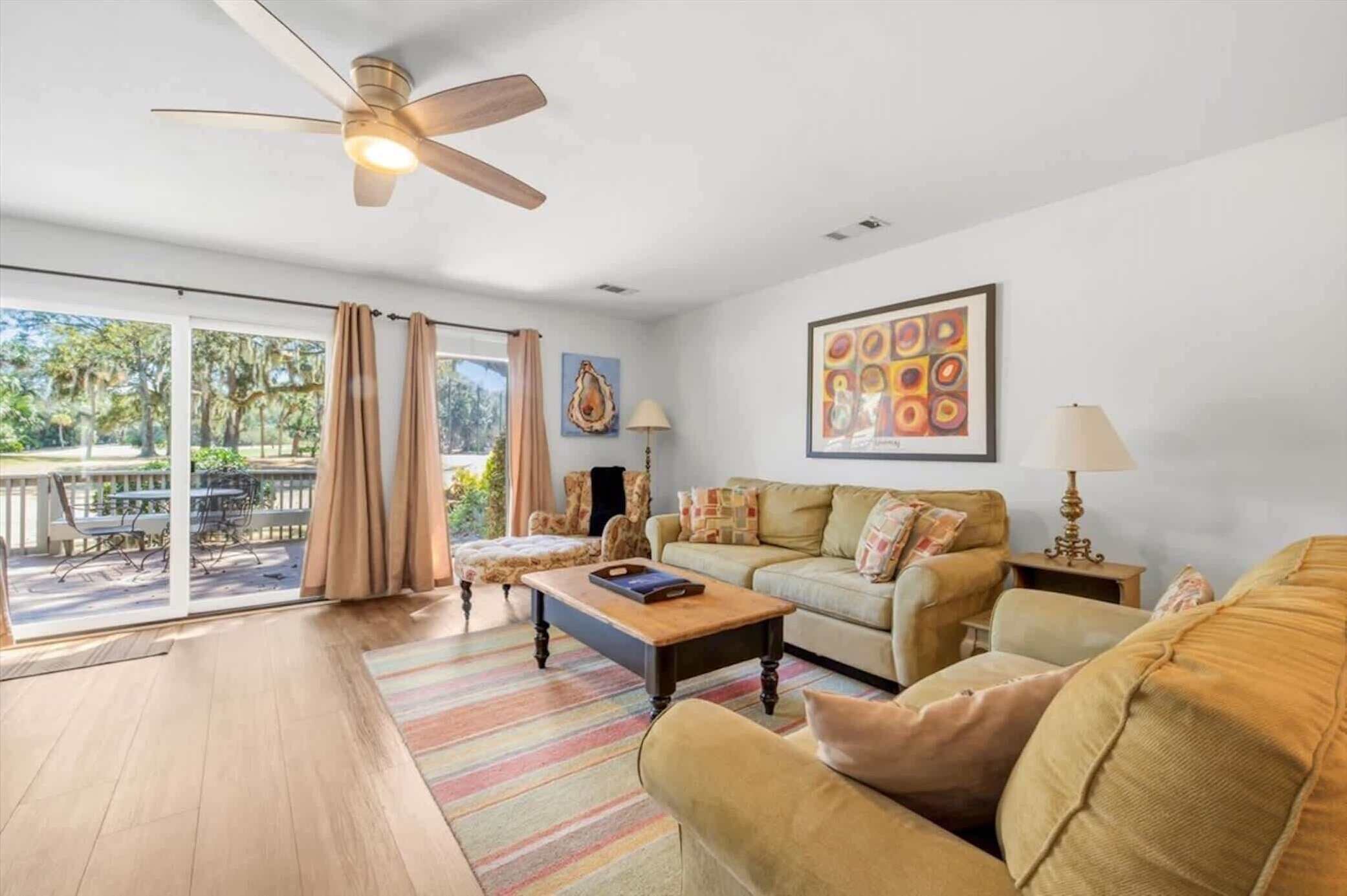 A spacious living room with a ceiling fan, a large window with curtains, a sofa set, a coffee table, and a painting on the wall.