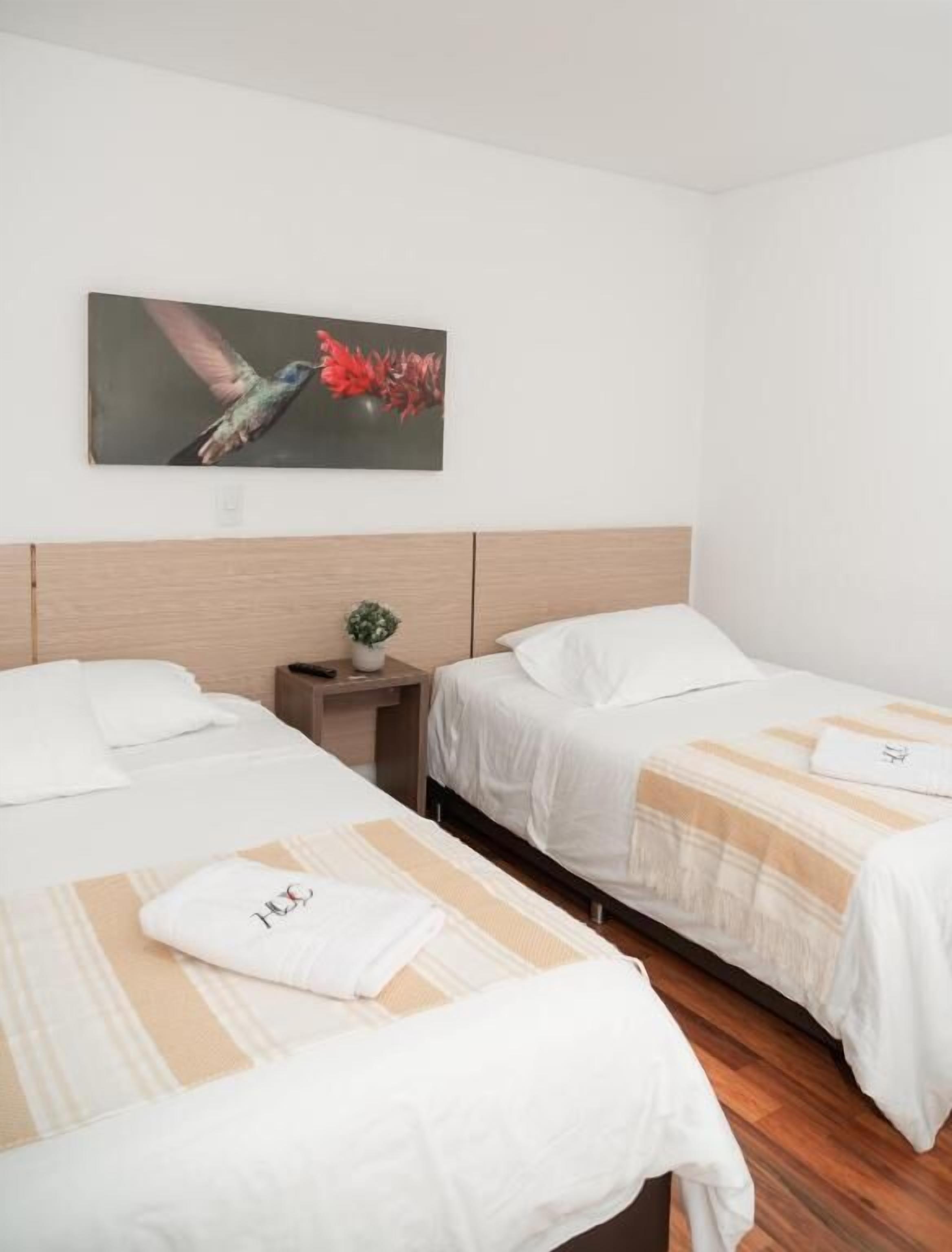 A hotel room with two single beds, a nightstand with a plant, and a painting of a hummingbird on the wall.