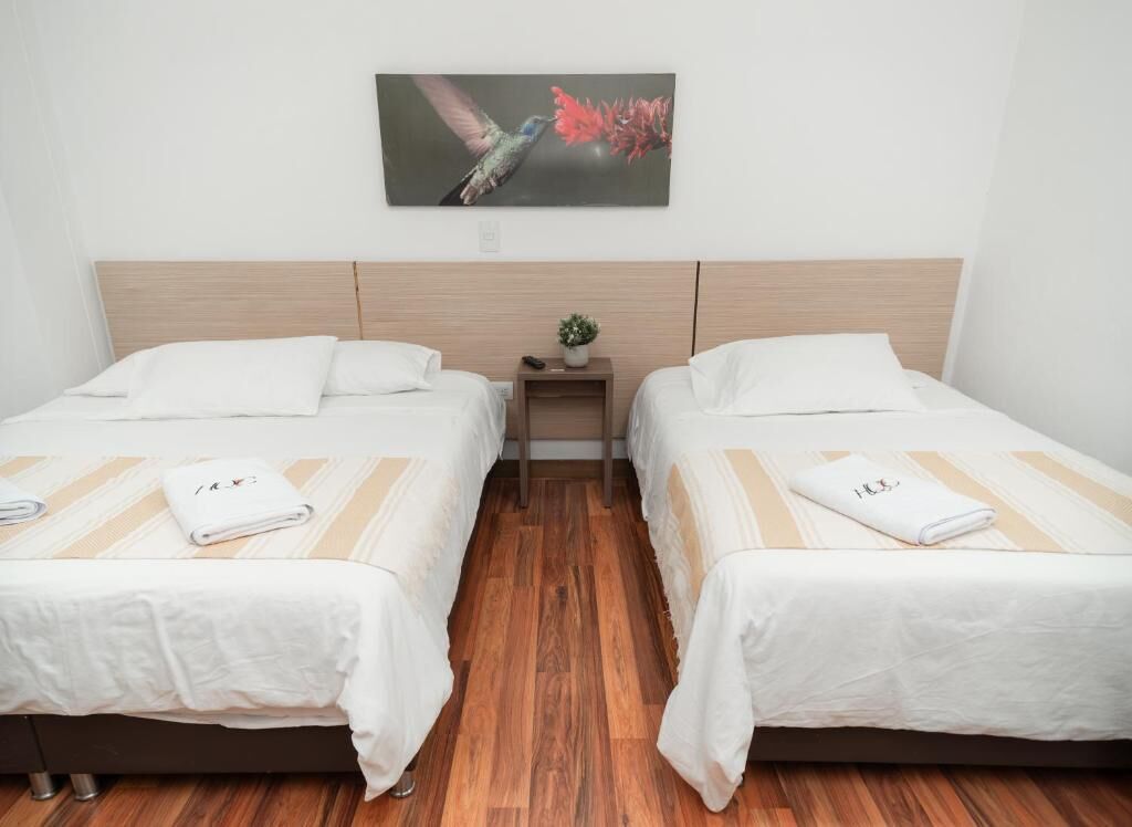 A hotel room with two beds, wooden flooring, a small bedside table with a plant, and a painting of a hummingbird on the wall.